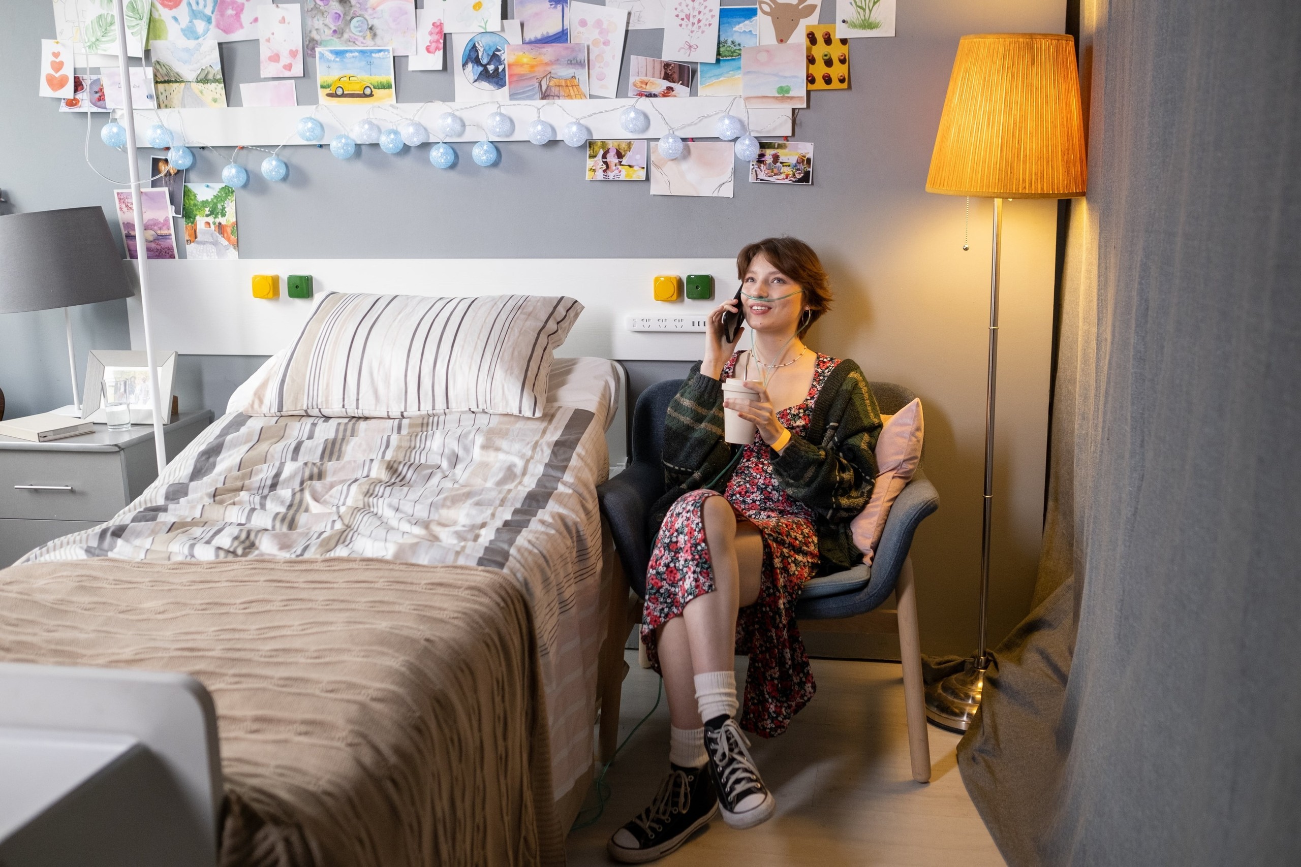 woman with breathing tube sitting  in a chair next to a bed while drinking from a coffee cup