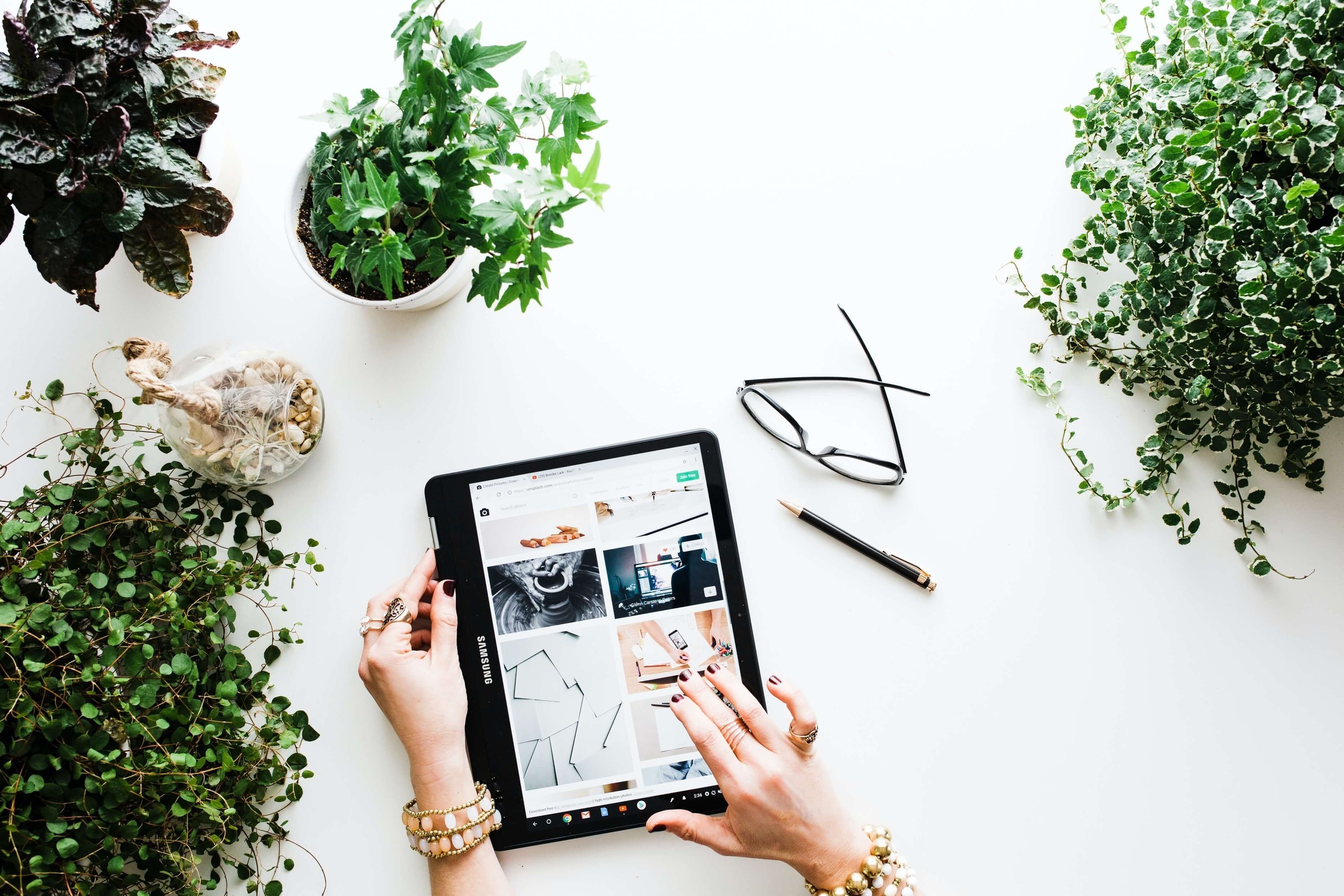 hands holding a tablet on top of table with a pen, glasses, and plants
